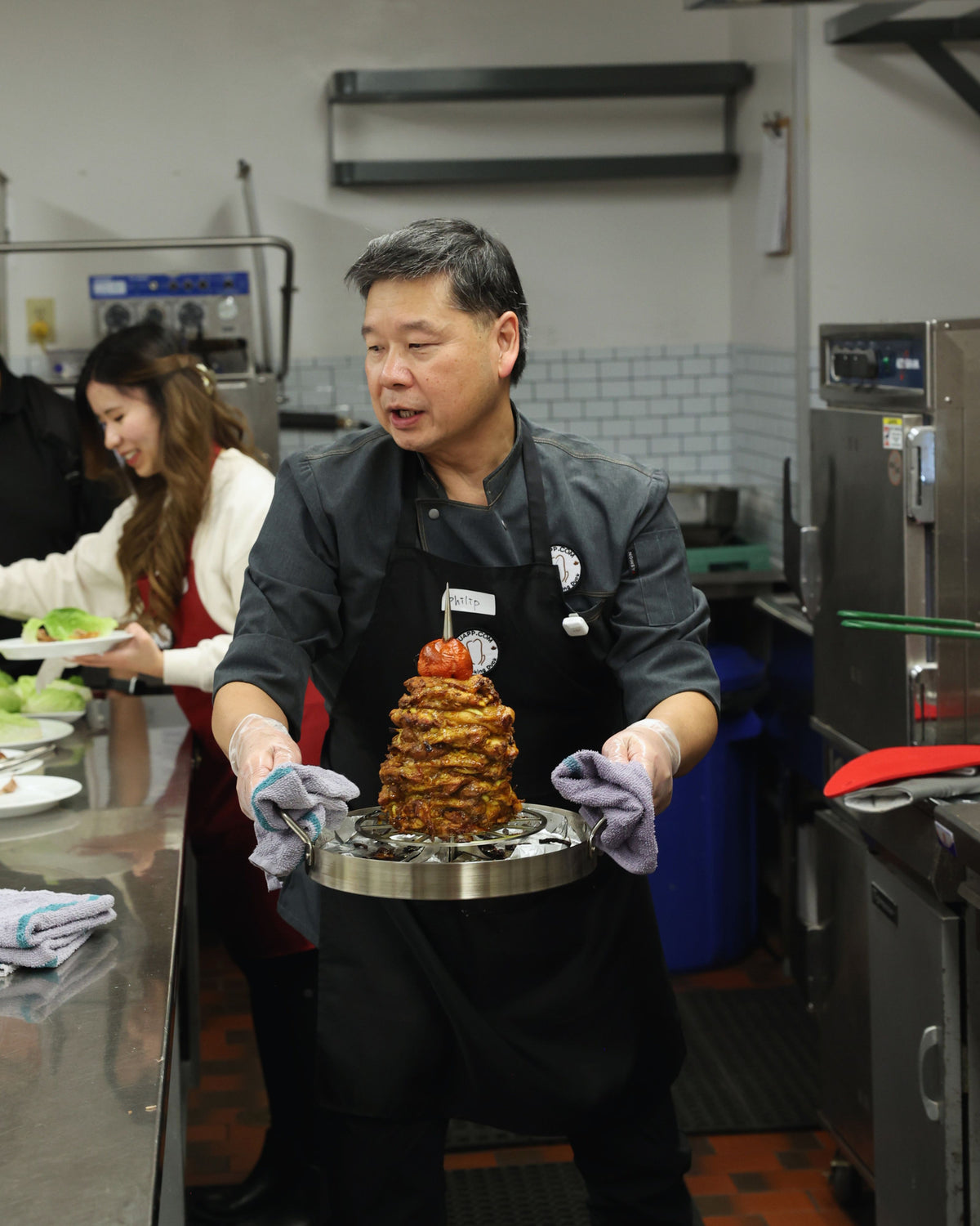 UpRoaster instructor carrying a finished vertical-roasted chicken shawarma stack from a DIY Peking Duck cooking class in a commercial kitchen.