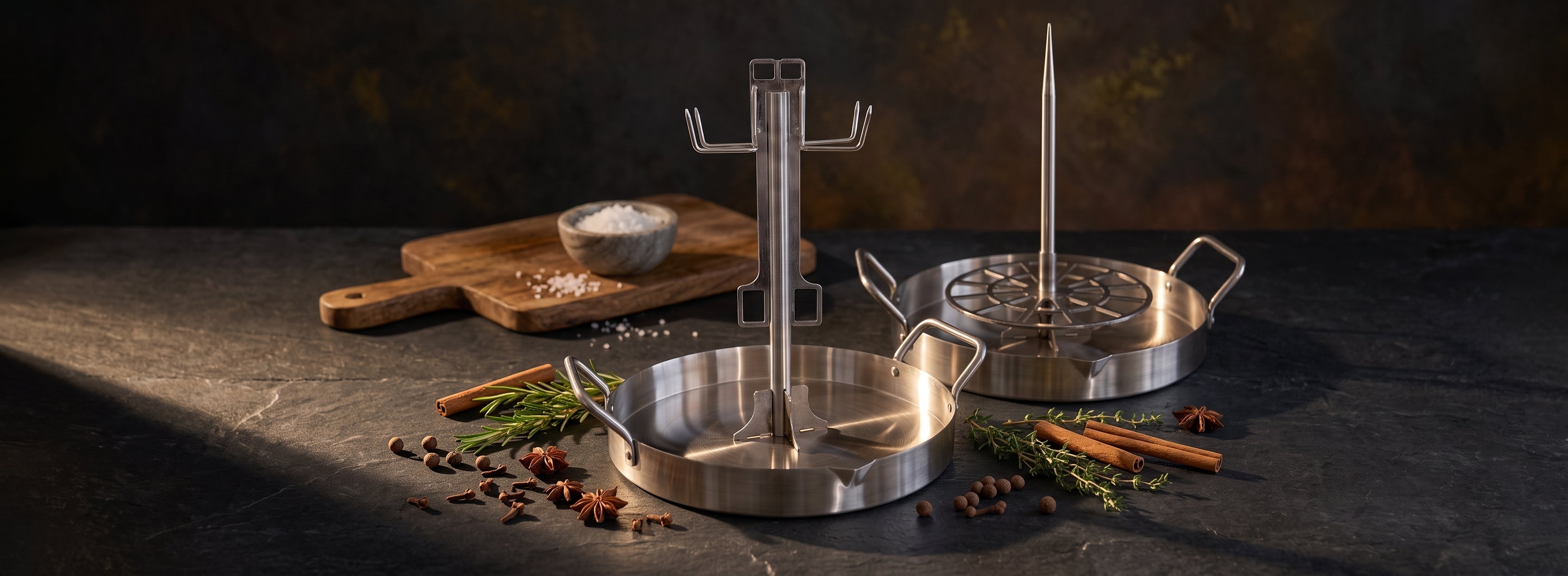 UpRoaster stainless steel vertical roasting pans with poultry hook and grill spit attachments, styled with fresh herbs and holiday spices on a dark countertop.
